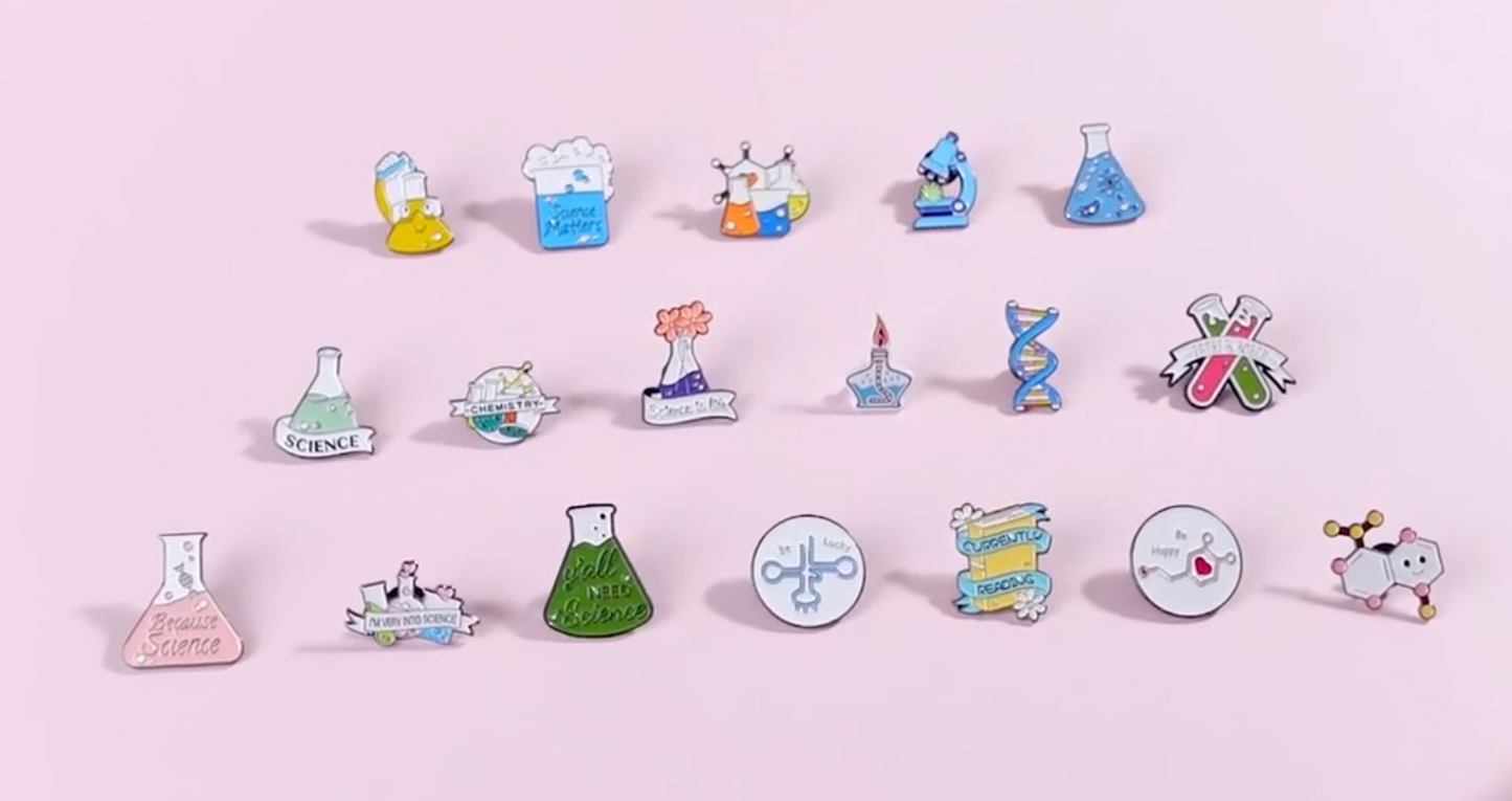 A set of colorful science-themed enamel pins arranged in three rows on a solid light pink background. The pins feature various scientific illustrations including beakers, flasks, microscopes, DNA helixes, and chemical symbols, with text such as 'Science' and 'Breathe Science'.