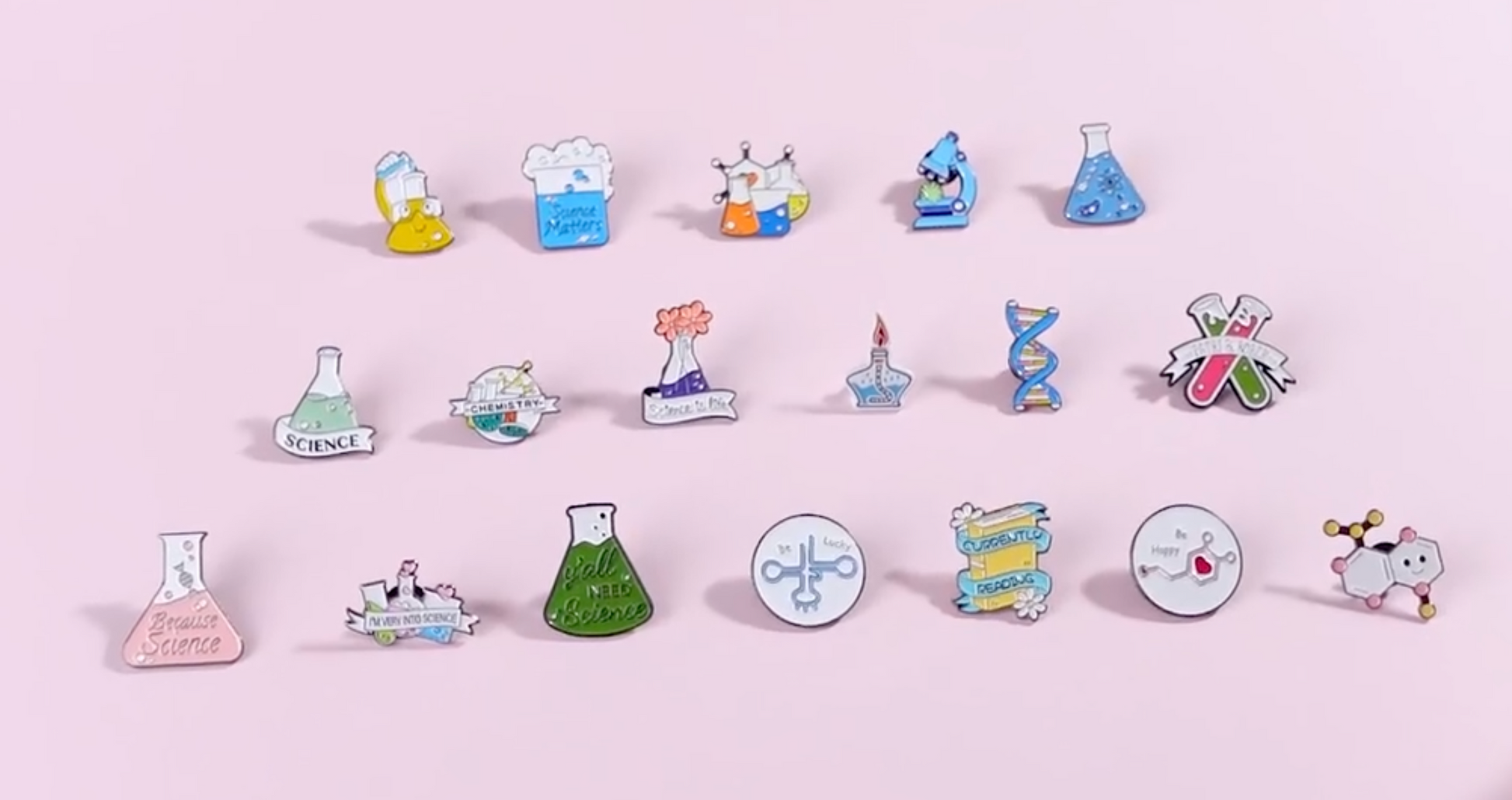 A set of colorful science-themed enamel pins arranged in three rows on a solid light pink background. The pins feature various scientific illustrations including beakers, flasks, microscopes, DNA helixes, and chemical symbols, with text such as 'Science' and 'Breathe Science'.