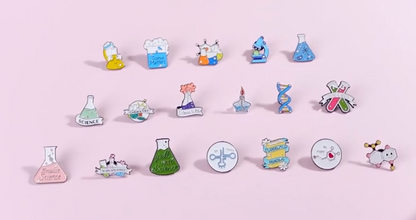 A set of colorful science-themed enamel pins arranged in three rows on a solid light pink background. The pins feature various scientific illustrations including beakers, flasks, microscopes, DNA helixes, and chemical symbols, with text such as 'Science' and 'Breathe Science'.