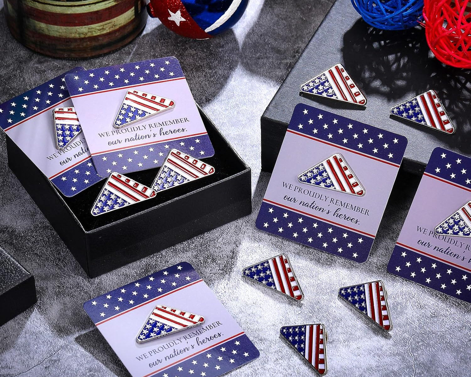 Patriotic-themed enamel pins and matching cards displayed in a black gift box on a gray textured surface, with decorative balls and woven balls in the background.